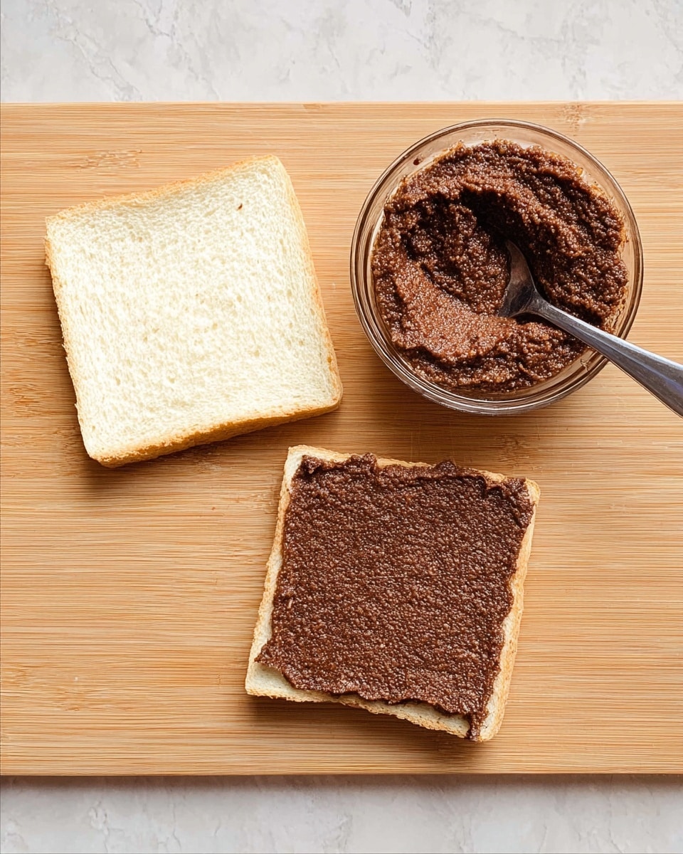 A top-down view shows two square slices of white bread placed on a light wooden surface; the slice on the left is plain, while the slice on the right is fully spread with a thick, dark brown, crumbly paste that has a slightly grainy texture. Above the slices, there is a small clear glass bowl filled with the same brown paste, with a shiny metal spoon resting inside it. The background is changed to a white marbled texture. photo taken with an iphone --ar 4:5 --v 7