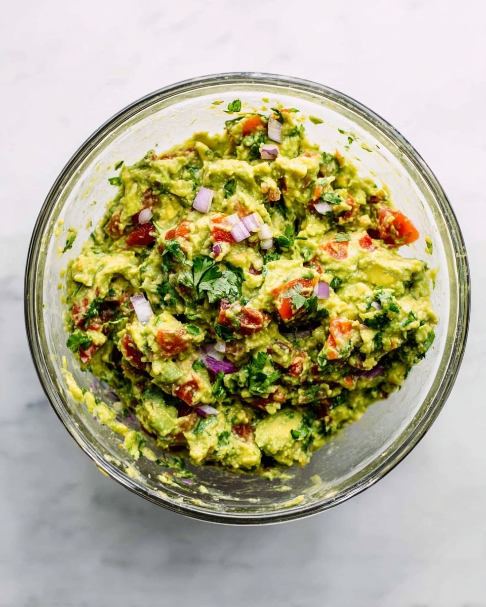 A clear glass bowl is filled with a fresh guacamole mix, consisting of mashed green avocado as the main layer, combined with small chunks of red tomato and bits of purple onion scattered evenly throughout. The guacamole also has finely chopped green cilantro spread over it, adding texture and color contrast. The bowl is placed on a white marbled surface, highlighting the vibrant colors of the guacamole photo taken with an iphone --ar 4:5 --v 7