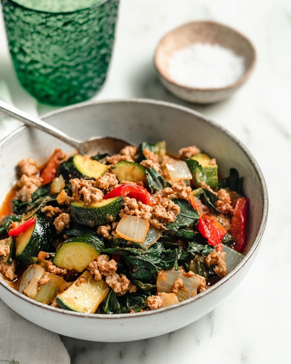 A white bowl filled with a colorful mix of cooked vegetables and ground meat. The bottom layer consists of dark green spinach leaves that look soft and slightly wilted. On top of the spinach are chunks of bright green zucchini, red bell pepper pieces, and white onion slices, all cooked with a slight shine from light sauce or oil. Ground meat in small, crumbly pieces mixes evenly with the vegetables, adding a light brown color. The bowl sits on a white marbled surface, next to a textured green glass and a small bowl of salt, both slightly blurred. A spoon is partially visible inside the bowl. Photo taken with an iphone --ar 4:5 --v 7
