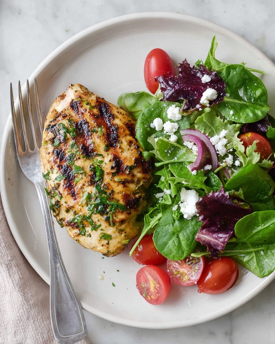 A white plate sits on a white marbled surface with a grilled chicken piece on the left side, showing clear dark grill marks and sprinkled with chopped green herbs. On the right side of the plate, there is a fresh salad made of green spinach leaves, mixed greens in shades of purple and green, and halved bright red cherry tomatoes. Small white crumbles of cheese are scattered over the salad. A silver fork rests on the edge of the plate on the left side. Photo taken with an iphone --ar 4:5 --v 7