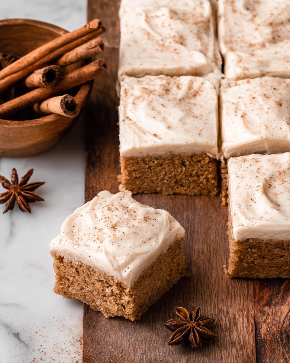 The image shows a thick square cake cut into nine pieces with a smooth, creamy frosting layer on top, sprinkled evenly with a light dusting of cinnamon giving it a speckled appearance; the cake layer underneath is moist and brown with a slightly crumbly texture. One piece is slightly lifted and tilted to reveal the cake’s soft, spongy inside beneath the white frosting. Next to the cake, on a wooden surface, there is a wooden bowl filled with cinnamon sticks and star anise. The entire scene is set against a white marbled texture surface. photo taken with an iphone --ar 4:5 --v 7