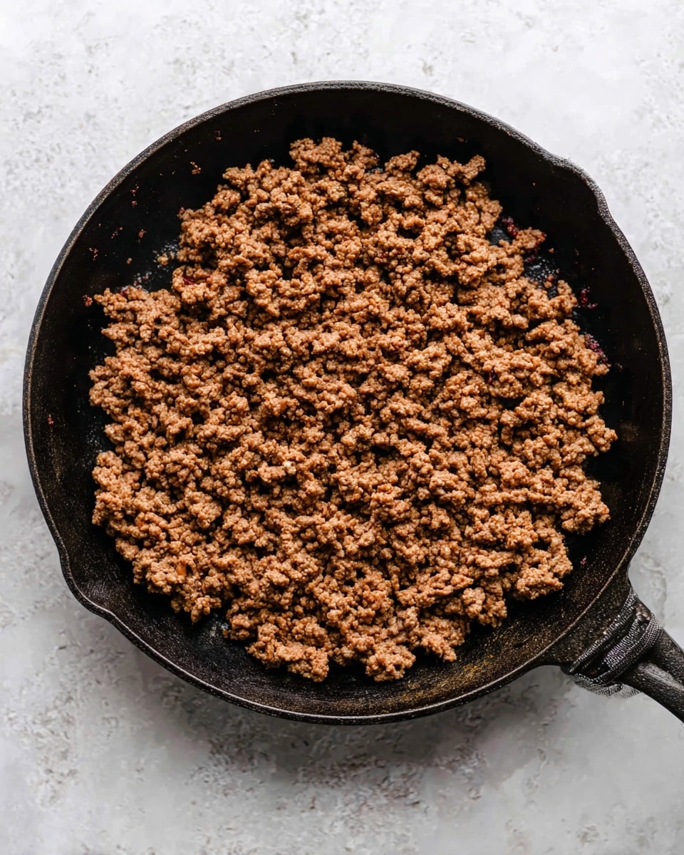 A close-up top view of a black cast iron skillet filled with browned ground meat spread evenly across the pan. The meat is crumbly with a mix of darker and lighter brown colors showing slight steam rising from it. The skillet sits on a white marbled surface, creating a clean and bright contrast against the dark pan and meat. The pan has a short handle visible on the right and a small lip on the left. photo taken with an iphone --ar 4:5 --v 7