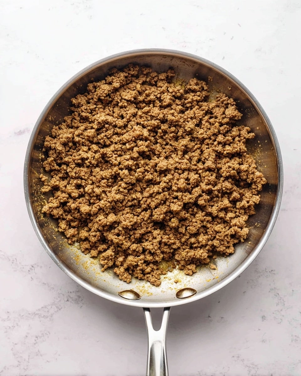 The image shows a silver metal pan with cooked ground meat inside. The meat is brown and crumbled with some small pieces, spread evenly across the bottom of the pan. The pan has a handle on the left side and a small metal loop on the right side. The background surface is a white marbled texture. photo taken with an iphone --ar 4:5 --v 7