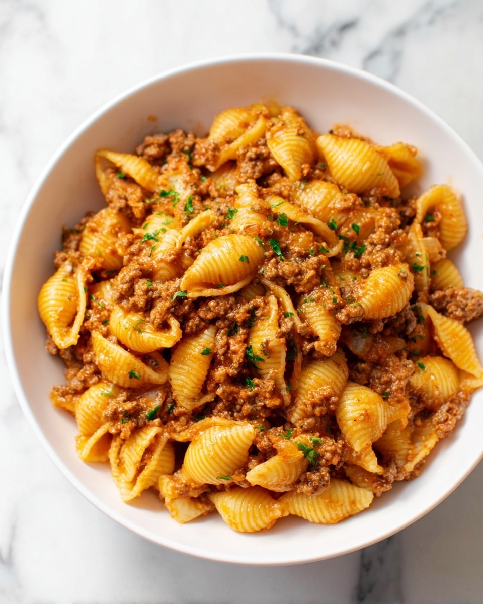 A white bowl filled with about two layers of shell pasta mixed with ground meat and small pieces of cooked onion. The pasta shells are yellow-orange and have a slightly shiny surface, coated evenly with a thick brownish-orange meat sauce that clings to both the pasta and meat. Small green herb bits are scattered on top and mixed in, giving some fresh color contrast. The bowl sits on a white marbled surface. photo taken with an iphone --ar 4:5 --v 7