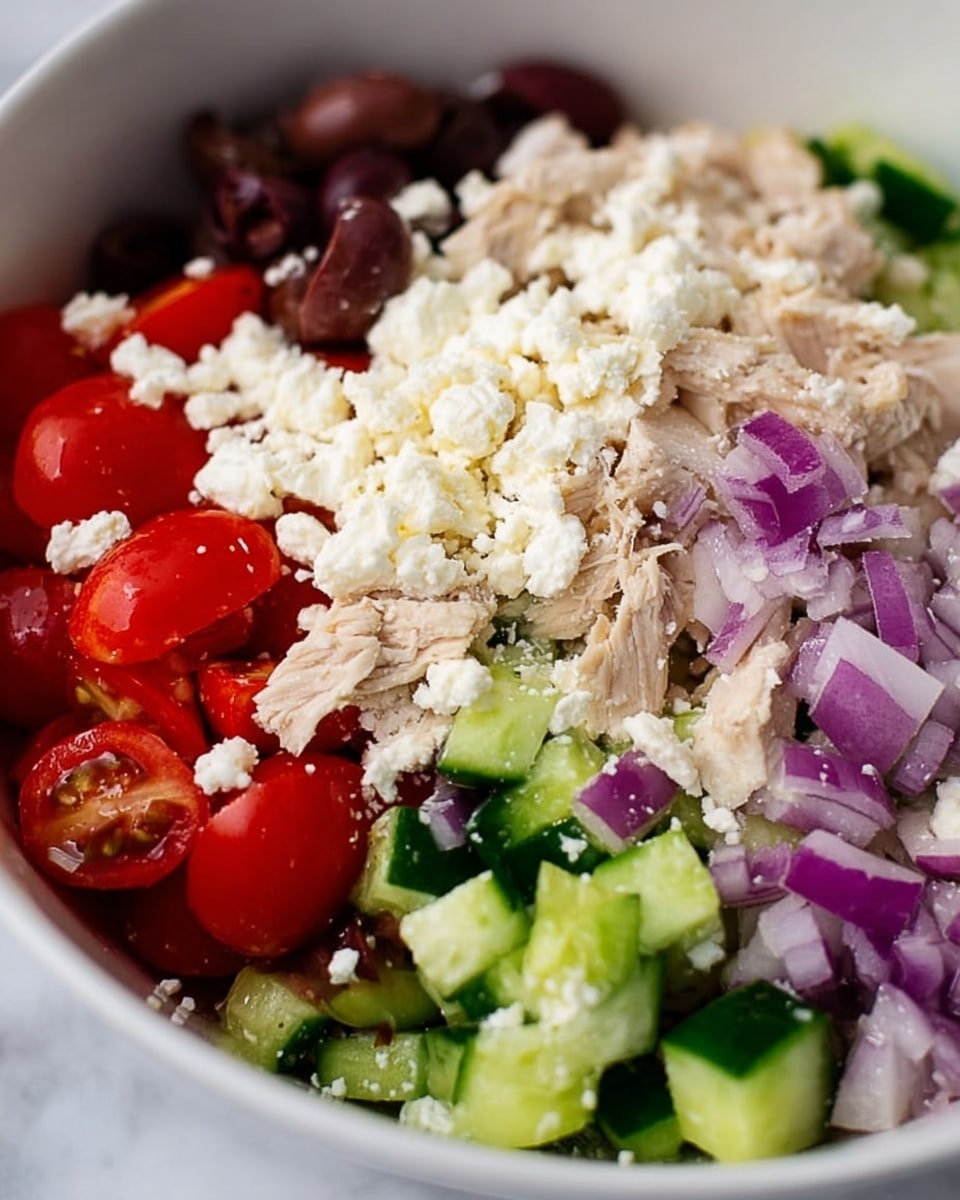 A close-up view of a fresh salad in a white bowl showing five layers of ingredients. The bottom layer has bright red cherry tomato halves, beside a layer of torn light beige cooked chicken pieces. Next is a layer of small green cucumber chunks, followed by creamy white crumbled cheese scattered all over. The top layer has chopped purple-red onions and sliced dark brown olives, all sitting on a white marbled surface. Photo taken with an iphone --ar 4:5 --v 7