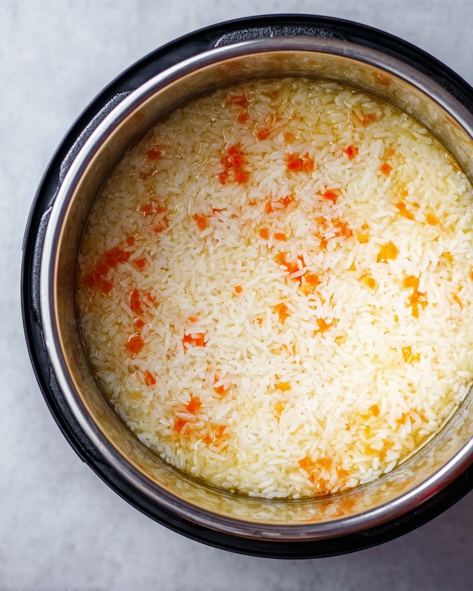 The image shows a close-up top view of an Instant Pot filled with a single layer of cooked rice mixed with small pieces of orange carrots. The rice is white and soft with a glossy, moist texture, while the carrot pieces are scattered evenly within the rice, adding bright pops of color. The Instant Pot has a shiny metallic inner surface and a black outer edge, sitting on a white marbled surface. The photo taken with an iphone --ar 4:5 --v 7