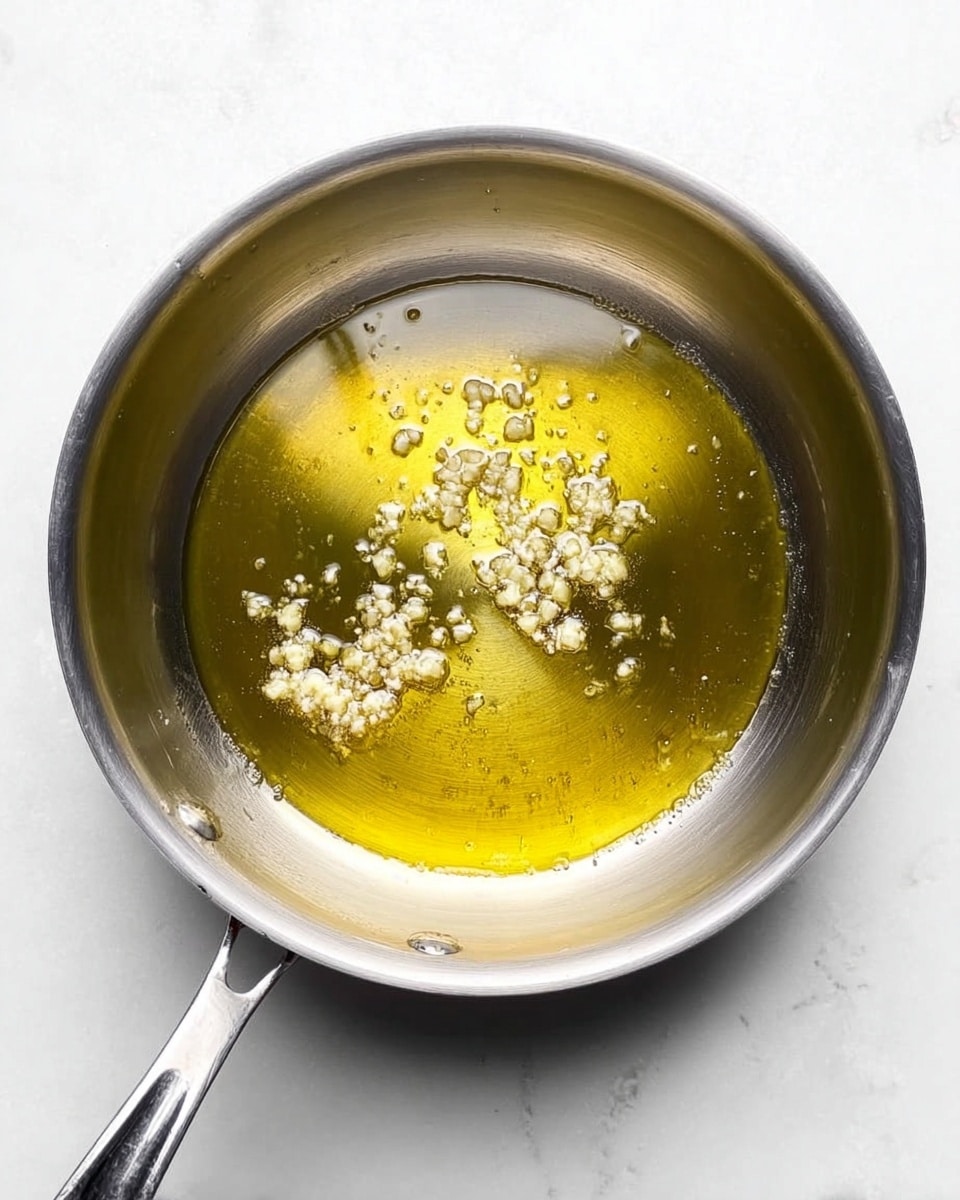 A shiny silver pot filled with a thin layer of warm golden oil sits on a white marbled surface. Small pieces of minced garlic float in the oil, creating clusters of light cream color spread unevenly around the center. The pot handle extends out to the left side, with a clear reflection of light showing the metal texture. The overall look is simple, clean, and fresh. photo taken with an iphone --ar 4:5 --v 7