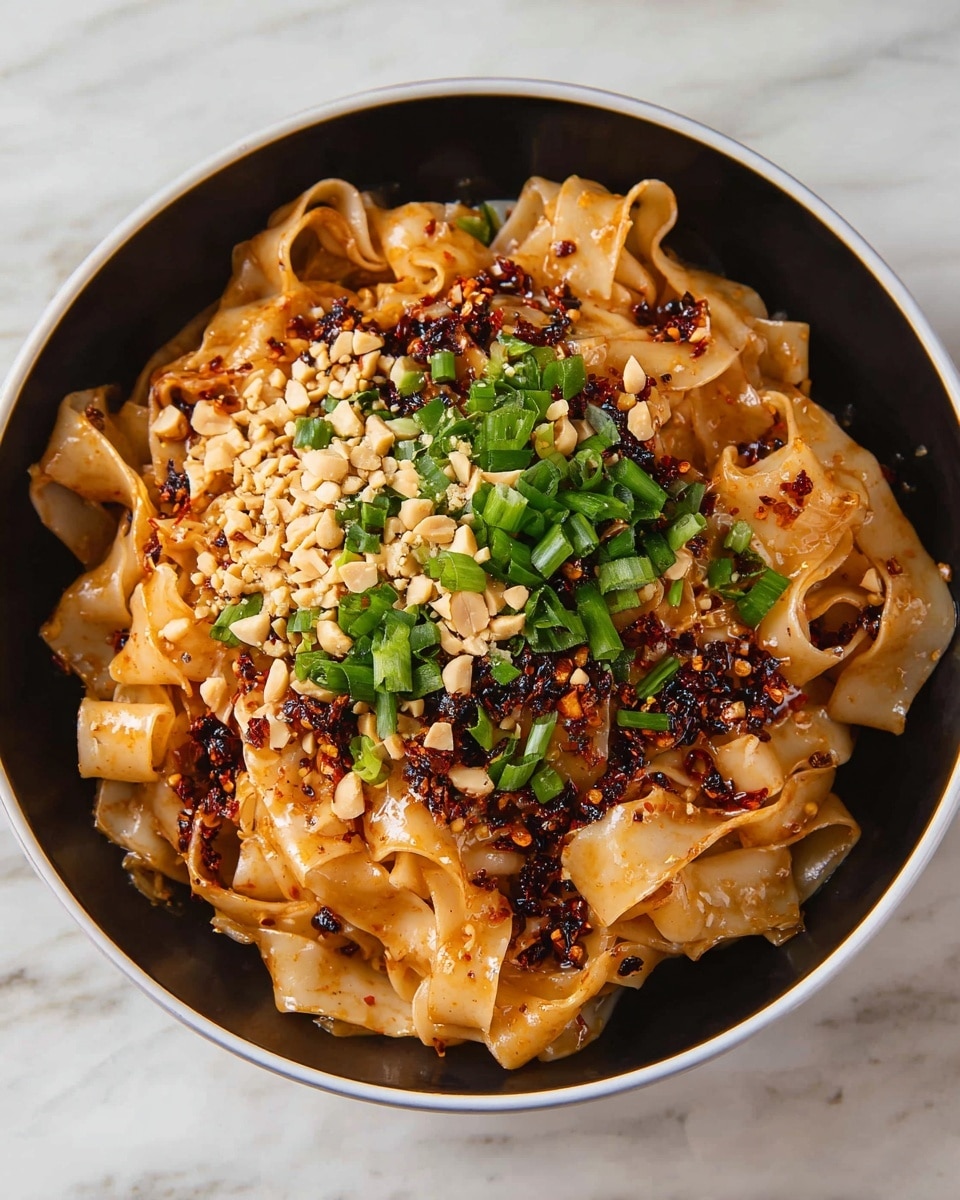 In a white bowl, there is a pile of wide, flat noodles that are light brown from a spicy sauce coating them. The noodles have a soft texture with some folds and curls. On top, there is a layer of finely chopped peanuts that are pale beige, scattered mostly in the center. Next to the peanuts, there is a bright green layer of sliced green onions, adding fresh color. Dark red chili flakes and oil are sprinkled over the noodles and toppings, adding contrast and shine. The bowl is placed on a white marbled surface. Photo taken with an iphone --ar 4:5 --v 7