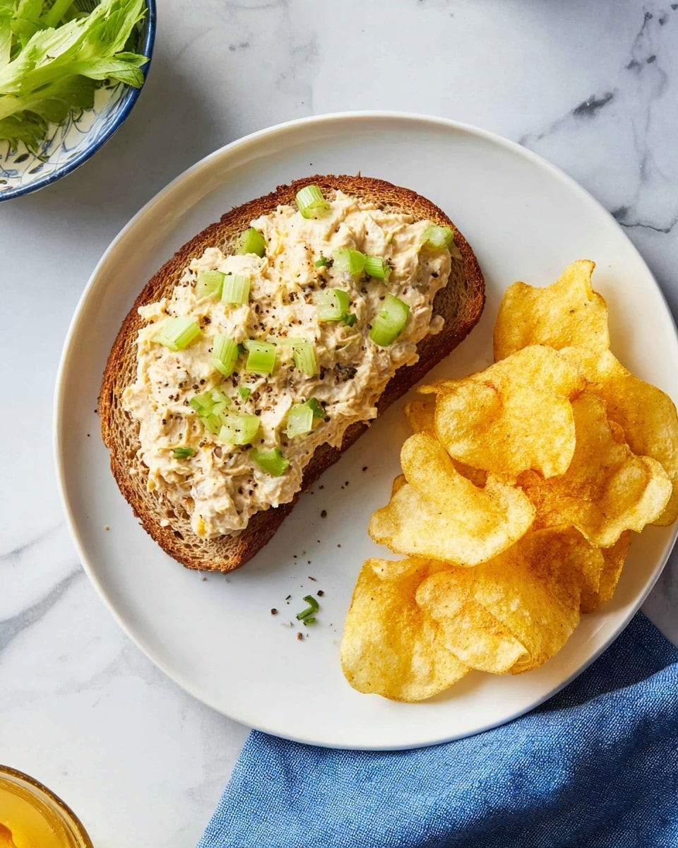 A white round plate sits on a white marbled surface, holding a slice of toasted brown bread topped with a creamy beige spread mixed with small green celery pieces and specks of black pepper. Next to the toast is a small pile of golden-yellow potato chips with a crunchy texture. Part of a blue cloth is visible at the bottom right corner, adding a soft contrast to the clean setting. Photo taken with an iphone --ar 4:5 --v 7