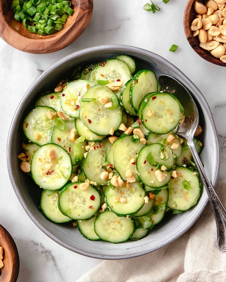A round gray bowl filled with one layer of thin cucumber slices, light green with dark green edges, each topped with small pieces of chopped peanuts and chopped green onions, scattered with a few red chili flakes. A silver spoon is inside the bowl resting on the cucumbers. On the white marbled surface around the bowl, there is a wooden bowl of chopped green onions on the top left and a wooden bowl of chopped peanuts on the top right, with some pieces scattered. A beige cloth is partially under the bowl on the right side. Photo taken with an iphone --ar 4:5 --v 7