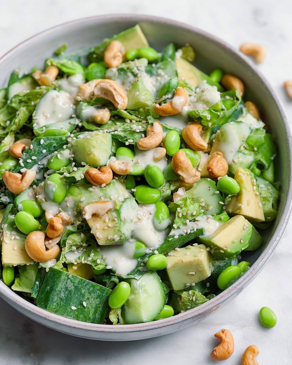 A close-up view of a fresh salad served in a white bowl, showing multiple layers of green vegetables including bright green edamame beans, light and dark green diced cucumbers, and chunks of pale green avocado, topped with crunchy golden brown cashew nuts and sprinkled with white sesame seeds. A light creamy dressing is drizzled unevenly over the salad, adding a smooth texture that contrasts with the fresh vegetables. The bowl sits on a white marbled surface with a few scattered sesame seeds around it. photo taken with an iphone --ar 4:5 --v 7