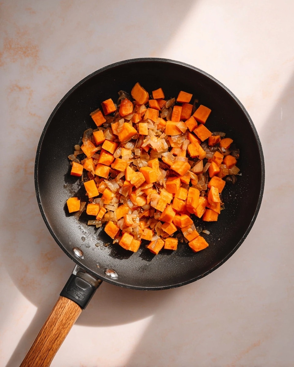 A black frying pan with a wooden handle holds small cubes of cooked bright orange sweet potatoes mixed with light brown sautéed onions. The pan sits on a white marbled surface with soft shadows around it. The sweet potatoes have a soft texture and warm color, evenly spread in the pan’s center. Photo taken with an iphone --ar 4:5 --v 7