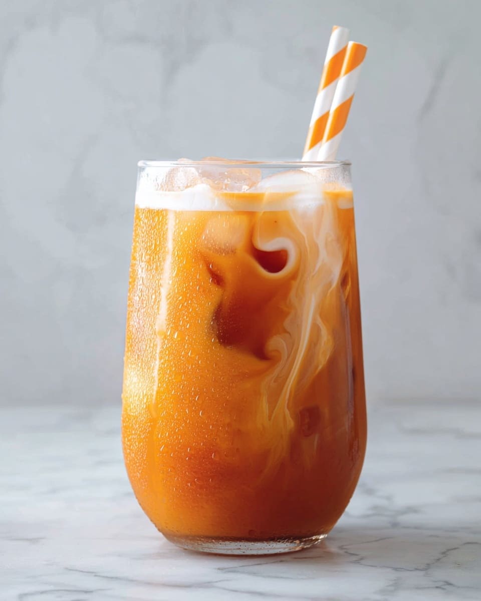 The image shows a clear glass filled with iced Thai tea. The drink has three visible layers: at the bottom, a dark orange tea layer; in the middle, the tea blends with cream creating a lighter orange swirl; and on the top, a creamy white layer mixing with ice cubes that are partly visible. Two straws stick out of the glass, one with white and orange stripes and the other plain white. The glass is set on a white marbled surface with a white marbled background, and condensation forms on the glass, showing its cold temperature. photo taken with an iphone --ar 4:5 --v 7