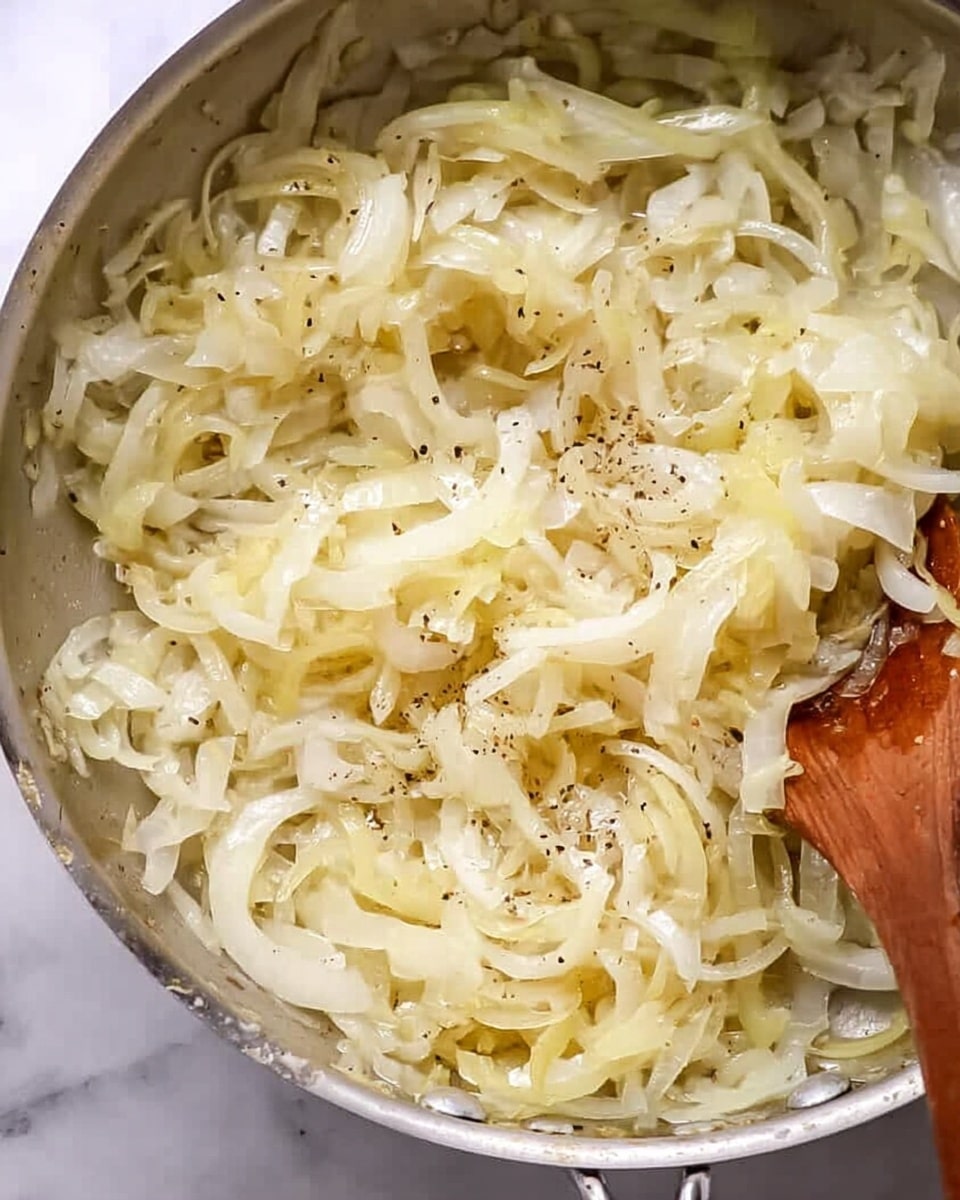 A close-up view of a silver pan filled with thinly sliced onions that are soft and cooked, showing a light yellow color with some pieces slightly translucent. The onions are seasoned with small specks of black pepper spread evenly across the surface. A wooden spoon is partially visible at the bottom right corner, stirring the onions. The background surface is a white marbled texture. photo taken with an iphone --ar 4:5 --v 7