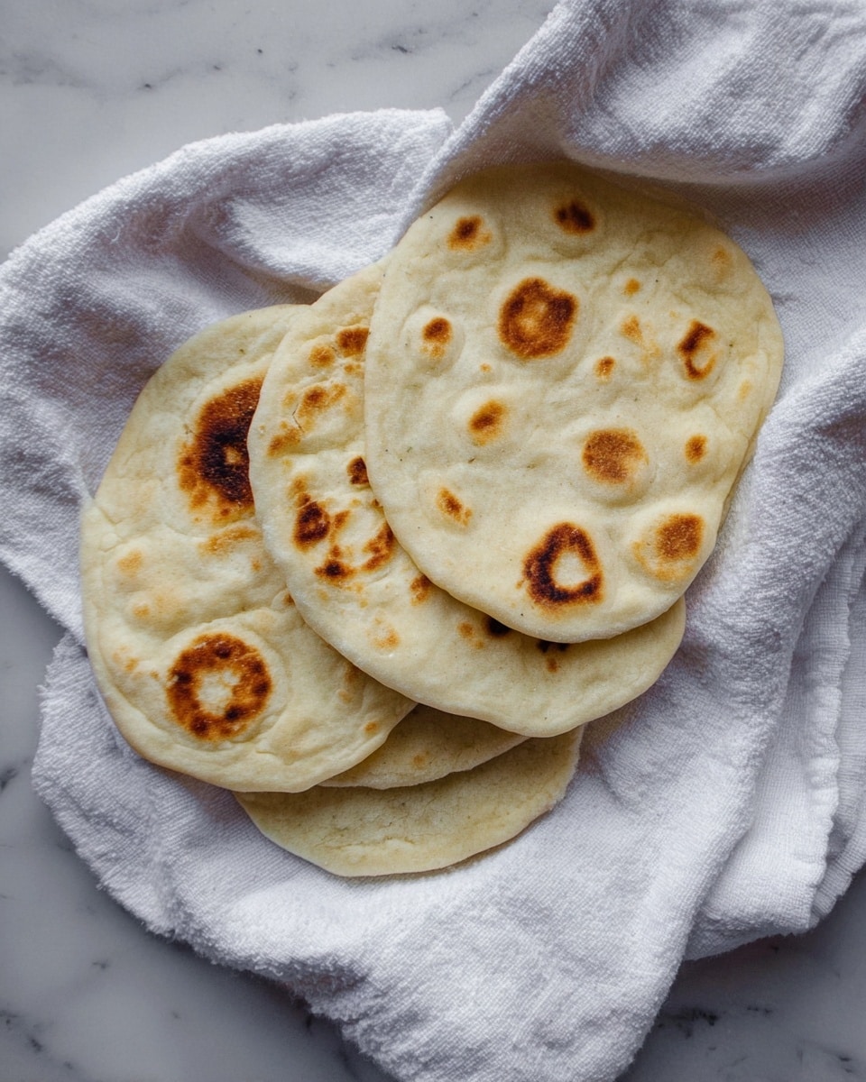 Three round flatbreads with light beige color and dark brown spots are stacked unevenly on a crumpled white cloth. The flatbreads have a soft, slightly puffed texture with uneven edges. The cloth is placed on a white marbled surface that adds subtle texture to the background. The overall look is simple and fresh, focusing on the warm tones and soft textures of the flatbreads and the cloth. photo taken with an iphone --ar 4:5 --v 7